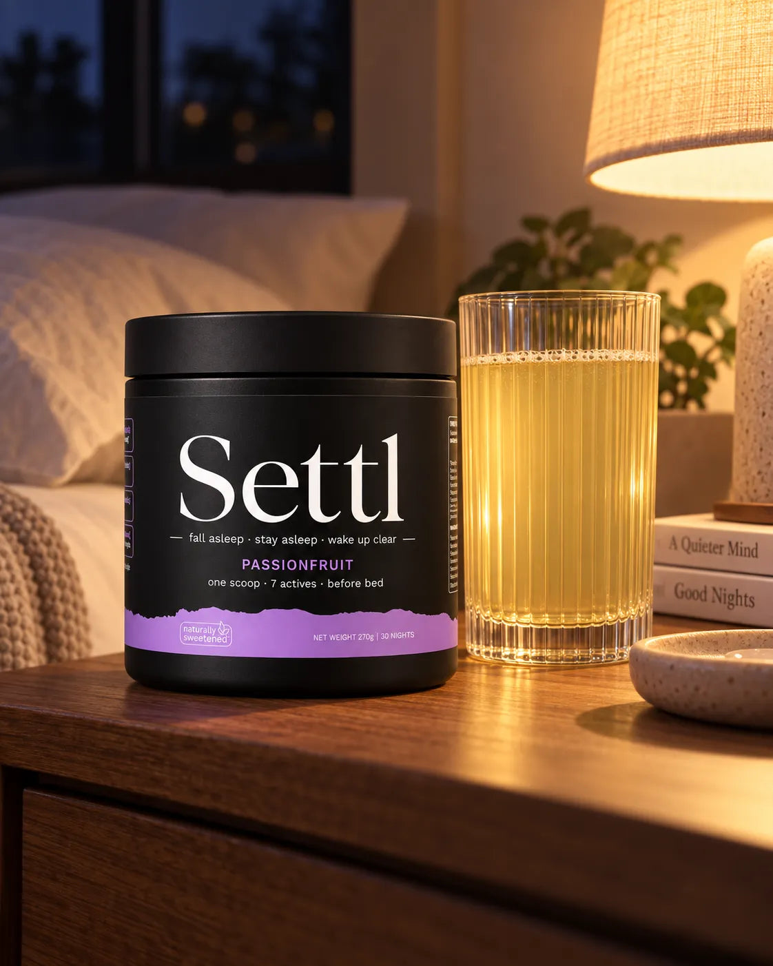 A tub of Settl Passionfruit and a glass of it on a bedside table under a warm lamp at night.