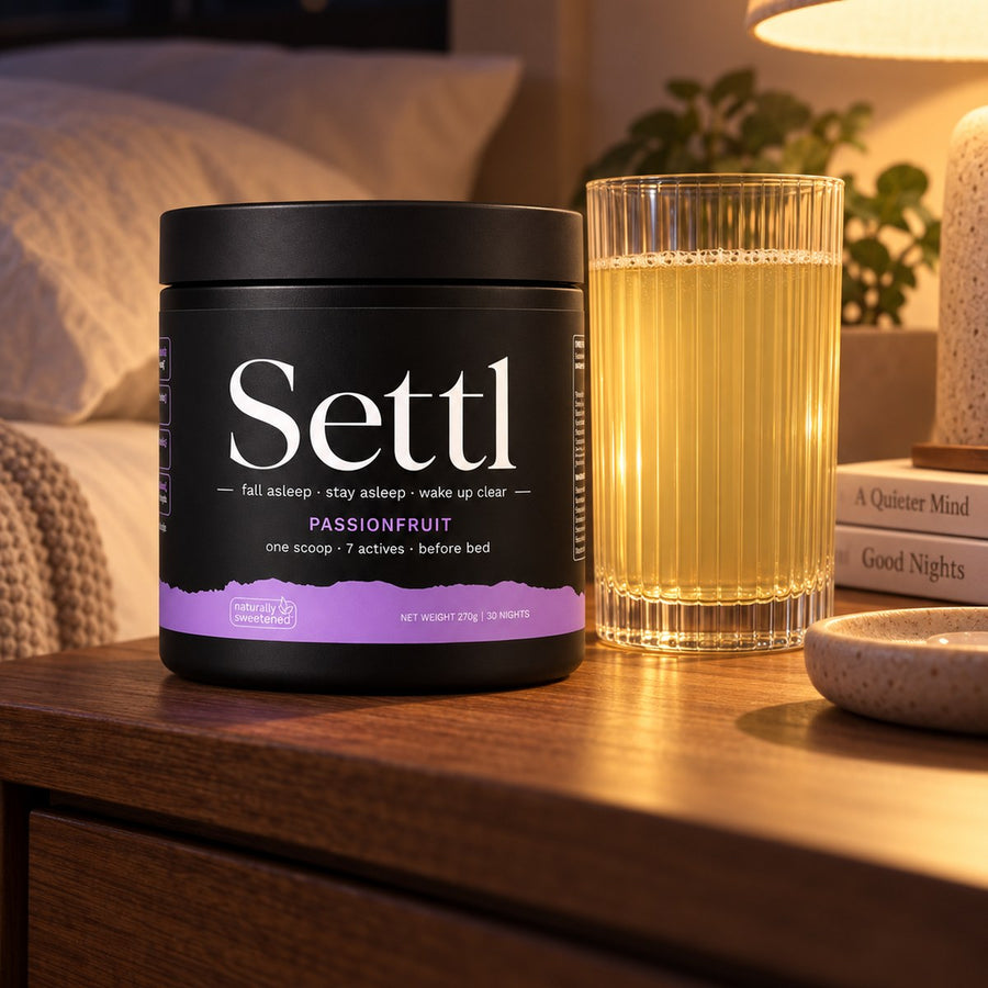A tub of Settl Passionfruit and a glass of the mixed drink on a bedside table, lit by a lamp at night