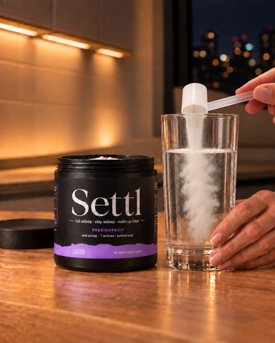 A scoop of Settl being tipped into a tall glass of cold water beside the tub, on a kitchen bench at night