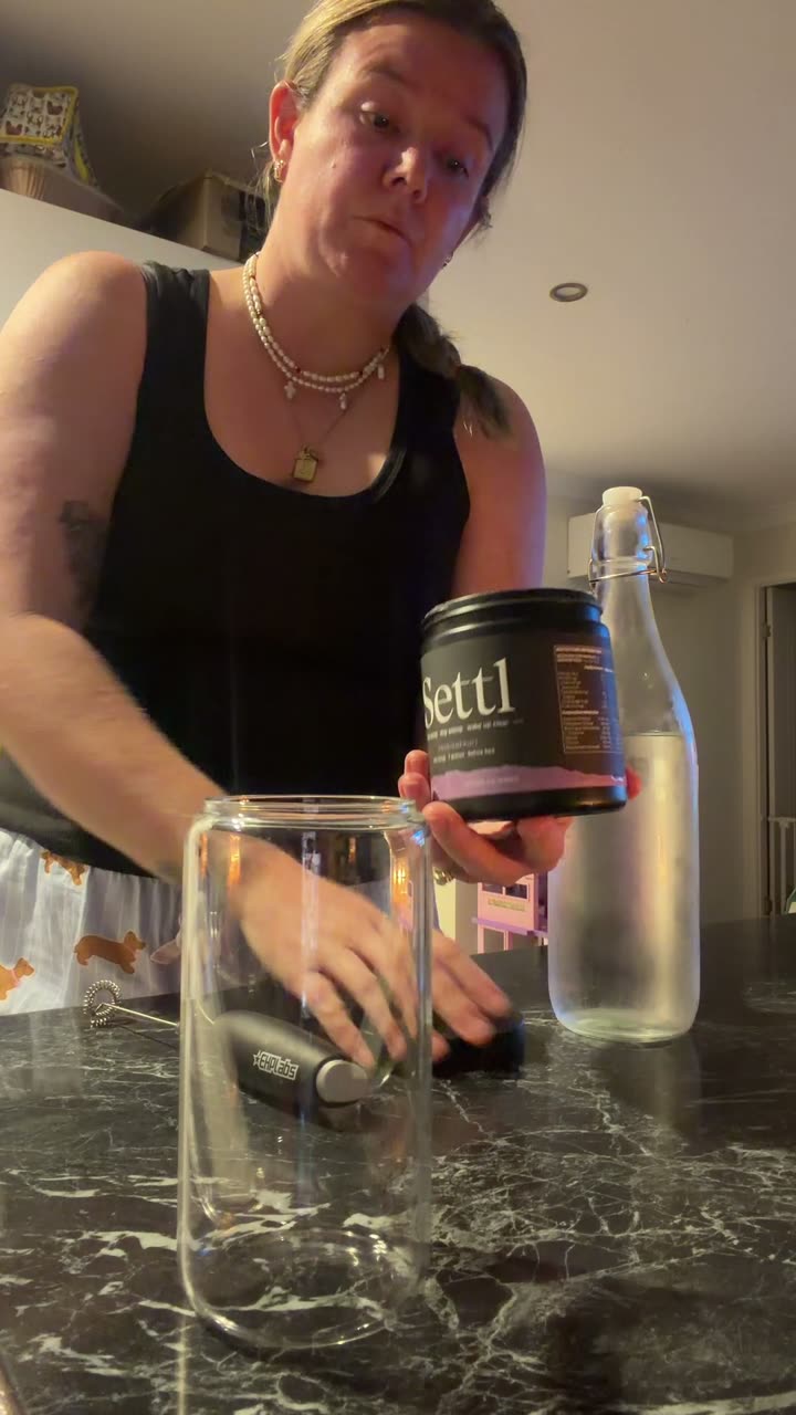Ashlie at her kitchen bench at night, holding a tub of Settl with an empty glass in front of her.